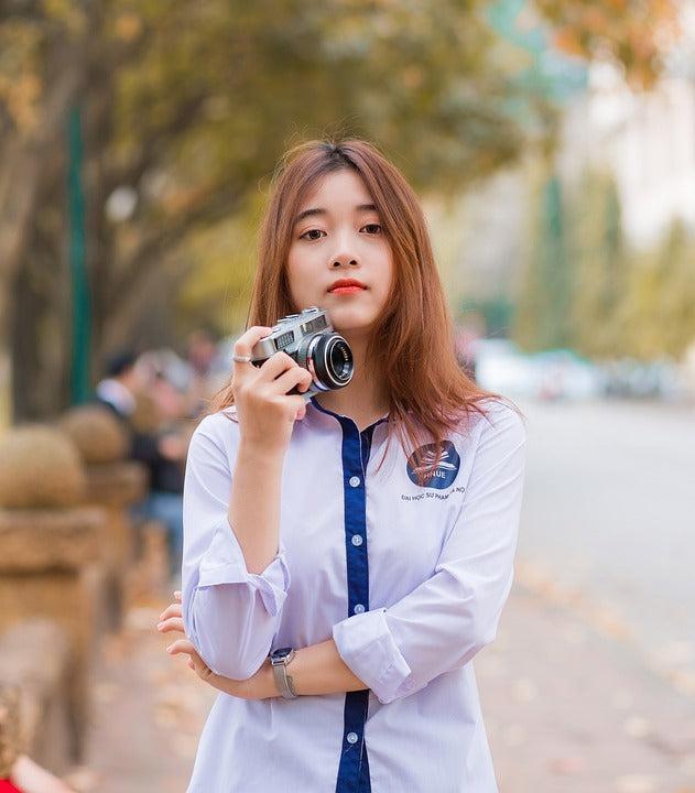 A young woman holding a camera stands confidently on a tree-lined street.